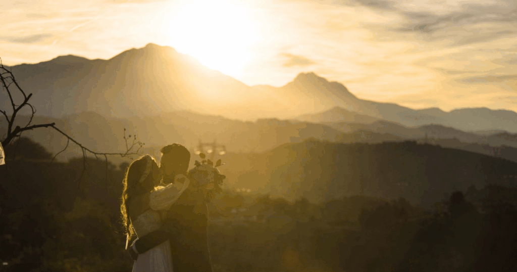 Coppia di sposi che si abbraccia e si bacia al tramonto, in controluce, con un panorama di montagne che rappresentano la Bella Addormentata sullo sfondo e una luce calda e dorata che avvolge la scena.