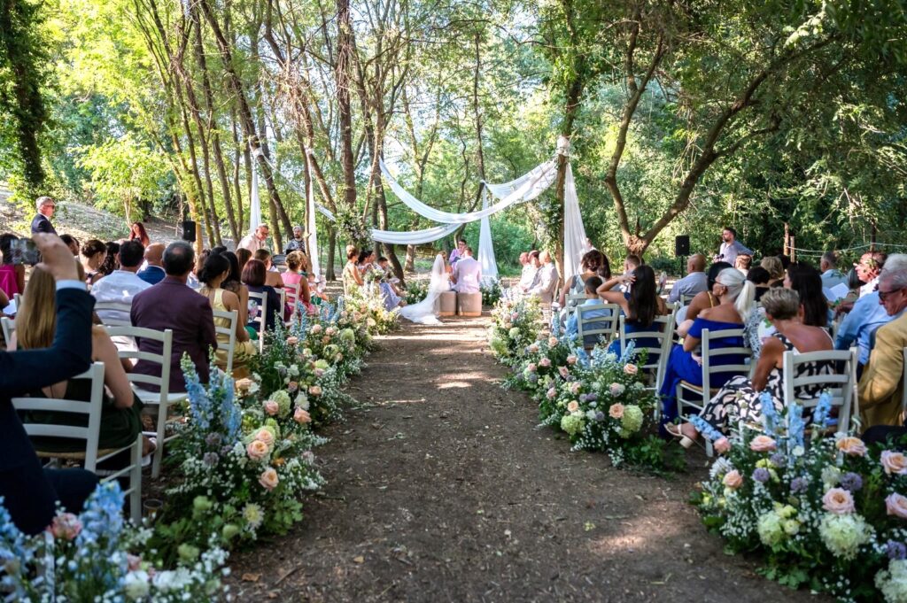 Cerimonia di matrimonio all’aperto in un bosco: gli sposi seduti davanti a un altare decorato con drappi bianchi e fiori, mentre gli invitati sono seduti su file di sedie ai lati di un corridoio ornato da composizioni floreali.